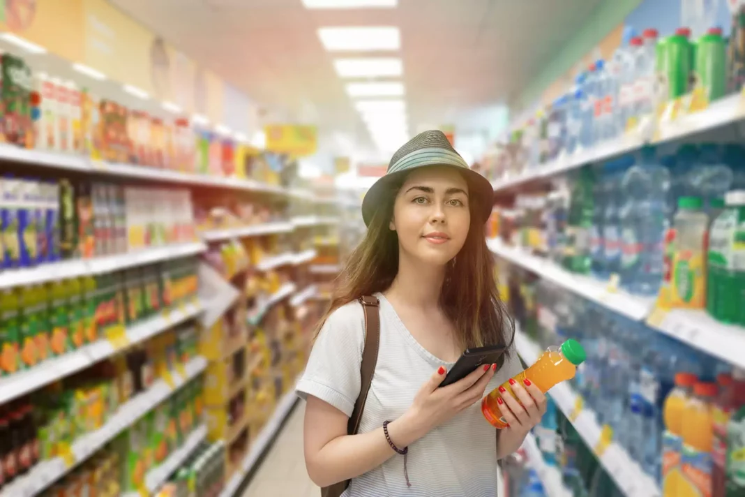 Young beautiful woman chooses a bottle of juice in the supermarket and checks the QR code on the label. The concept of modern technology and shopping in the store. Light. © STEKLO/ shutterstock.com